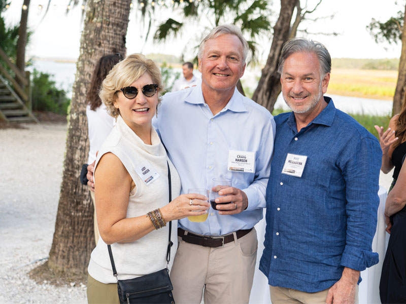 Three attendees at an outdoor waterfront reception, smiling and holding drinks under palm trees.